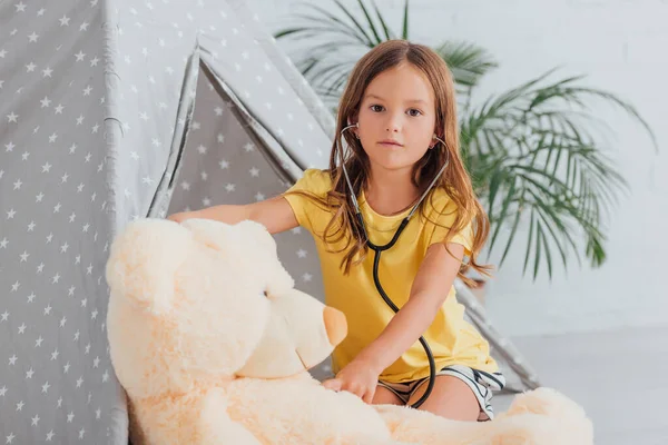 Kid in yellow t-shirt  looking at camera while playing doctor with teddy bear and stethoscope near kids wigwam — Stock Photo