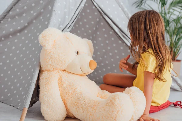 Girl in yellow t-shirt talking to teddy bear while playing near kids wigwam — Stock Photo