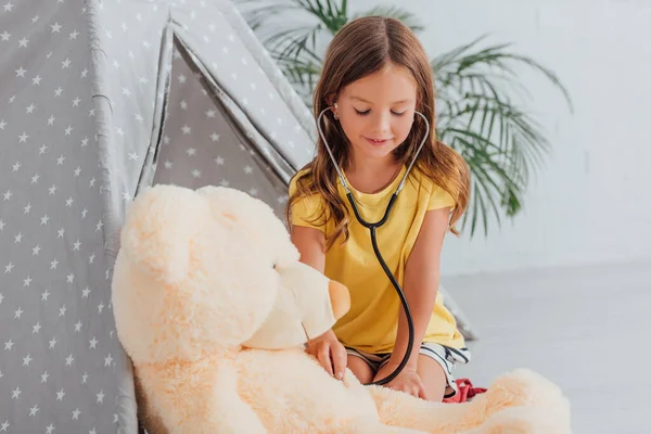 Girl in yellow t-shirt playing doctor with stethoscope and teddy bear near play tent — Stock Photo