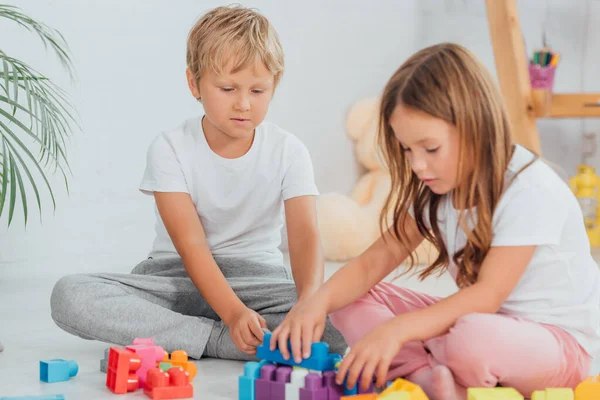 Focused brother an sister in pajamas sitting on floor and playing with building blocks — Stock Photo
