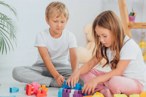 Irmão focado uma irmã de pijama sentado no chão e brincando com blocos de construção — Fotografia de Stock