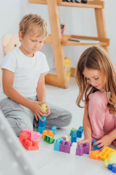 Селективное внимание детей в пижаме, сидящих на полу и играющих со строительными блоками — стоковое фото