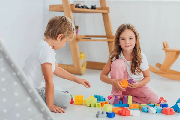 Enfoque selectivo de la chica mirando a la cámara mientras juega con bloques de construcción en el piso con el hermano - foto de stock