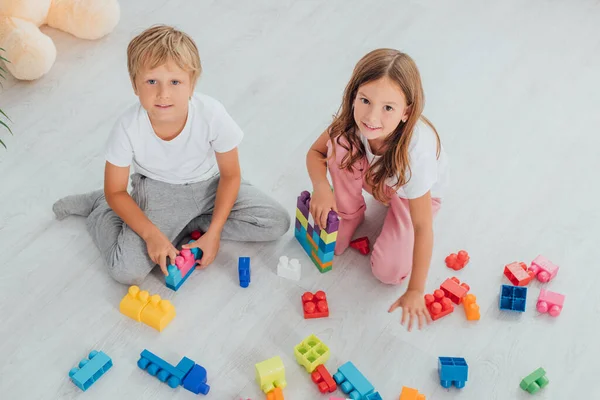 Vue grand angle des enfants en pyjama assis sur le sol près des blocs de construction et regardant la caméra — Photo de stock