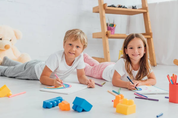 Bruder und Schwester im Schlafanzug blicken in die Kamera, während sie in der Nähe von Bauklötzen auf dem Boden liegen und mit Filzstiften zeichnen — Stockfoto