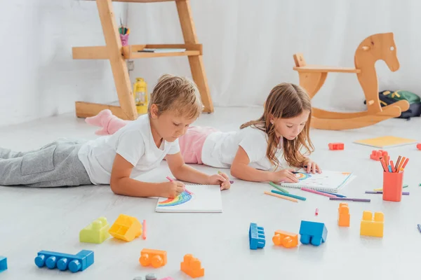 Hermano y hermana en pijama tendidos en el suelo cerca de bloques de construcción y dibujo con plumas de fieltro - foto de stock