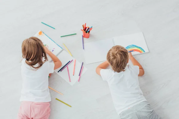 Top view of brother and sister in pajamas lying on floor and drawing with felt pens in sketchbooks — Stock Photo