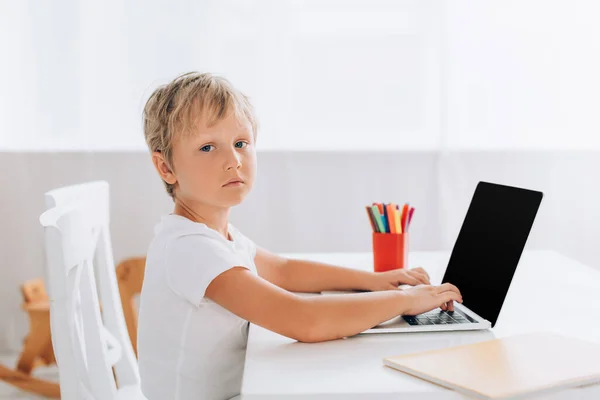 Menino sério em t-shirt branca olhando para a câmera enquanto sentado à mesa e usando laptop — Fotografia de Stock