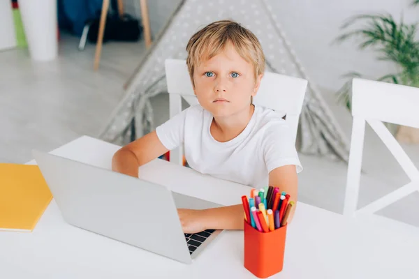 Hochwinkel-Ansicht des Jungen in weißem T-Shirt mit Laptop, während er am Tisch in der Nähe von Stifthalter und Kinder Pergwam auf dem Hintergrund sitzt — Stockfoto
