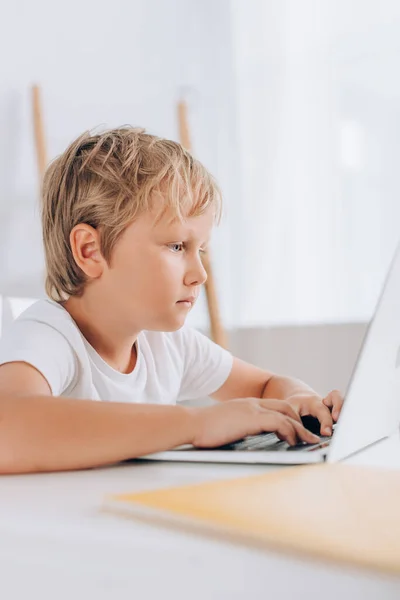 Enfoque selectivo de niño enfocado en camiseta blanca usando el ordenador portátil mientras está sentado en la mesa - foto de stock