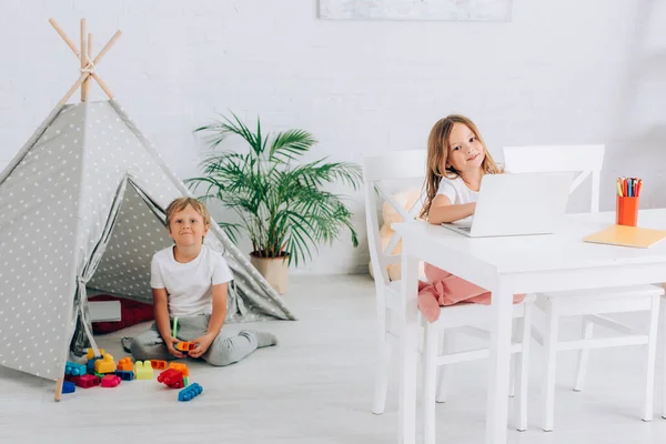 Mädchen mit Laptop am Tisch neben Bruder sitzt in der Nähe von Kindern Wigwam und Bausteine — Stockfoto