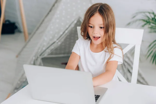 Vista de ángulo alto de la niña en camiseta blanca usando el ordenador portátil mientras está sentado en la mesa cerca de los niños wigwam - foto de stock