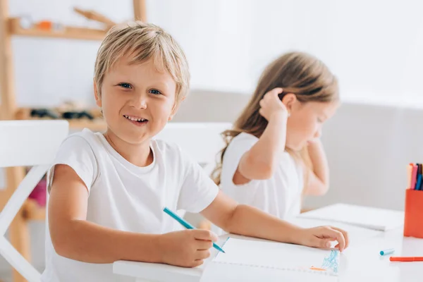 Aufgeregter Junge hält Filzstift in der Hand und blickt in die Kamera, während er neben Schwester am Tisch sitzt — Stockfoto