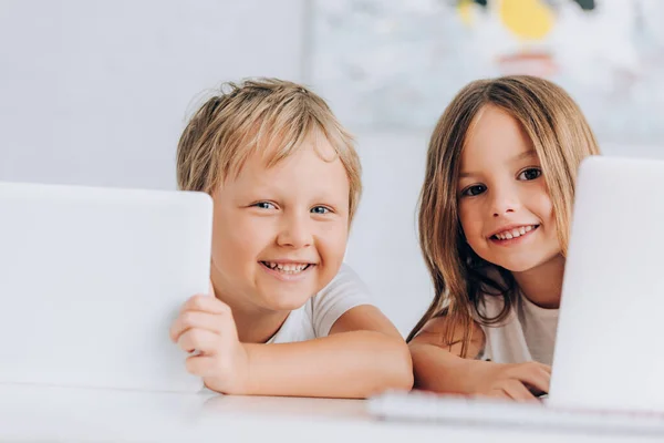 Aufgeregtes Geschwisterpaar blickt in die Kamera, während es neben Laptops sitzt — Stockfoto