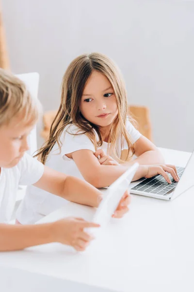 Foco seletivo da menina olhando para o irmão enquanto se sentam juntos à mesa e usando laptops — Fotografia de Stock