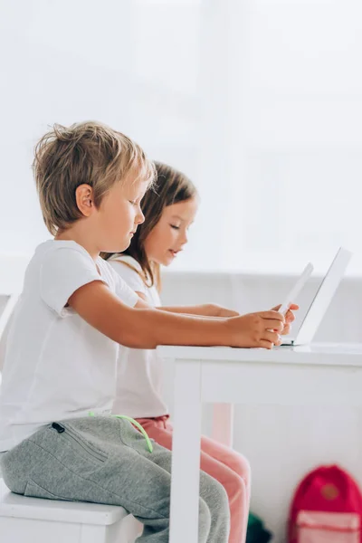 Vista lateral del hermano y la hermana en pijama sentados en la mesa y el uso de computadoras portátiles juntos - foto de stock