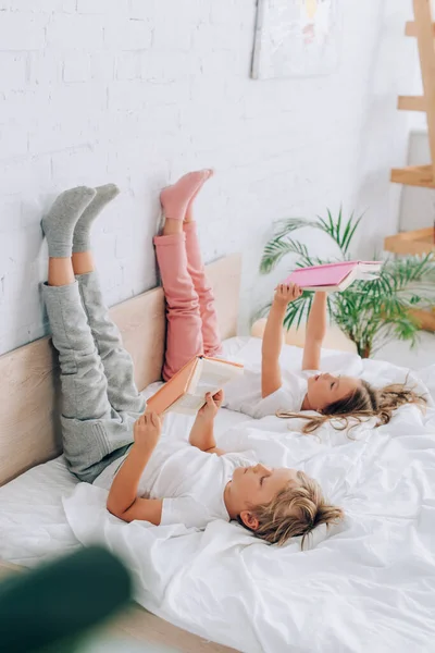 Selective focus of brother and sister in pajamas lying on bed with books and legs on wall — Stock Photo