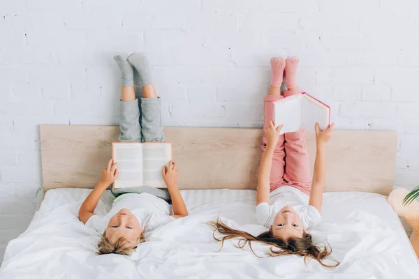 Irmão e irmã de pijama olhando para a câmera enquanto deitado com as pernas na parede e segurando livros — Fotografia de Stock