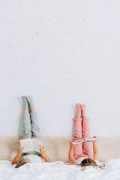 Frère et sœur en pyjama lisant des livres allongés avec les jambes sur le mur — Photo de stock