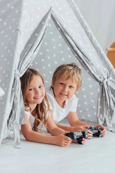 KYIV, UCRANIA - 21 de julio de 2020: hermano y hermana mirando a la cámara mientras yacen en los niños wigwam con joysticks - foto de stock