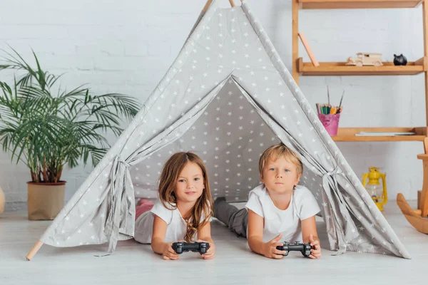 KYIV, UCRANIA - 21 DE JULIO DE 2020: hermano y hermana en pijama tumbados en el suelo en los niños wigwam y jugando videojuegos - foto de stock