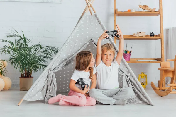 KIEW, UKRAINE - 21. JULI 2020: Junge zeigt Siegergeste in der Nähe von Schwester, die Steuerknüppel hält, während er auf dem Boden in der Nähe von Kindern sitzt — Stockfoto