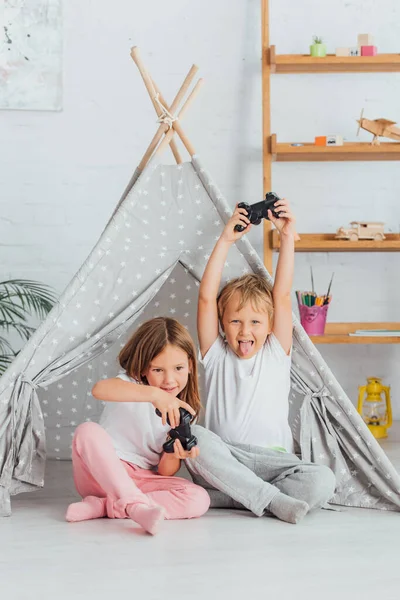 KYIV, UCRANIA - 21 de julio de 2020: niño emocionado sacando la lengua y mostrando el gesto ganador cerca de su hermana jugando videojuego - foto de stock