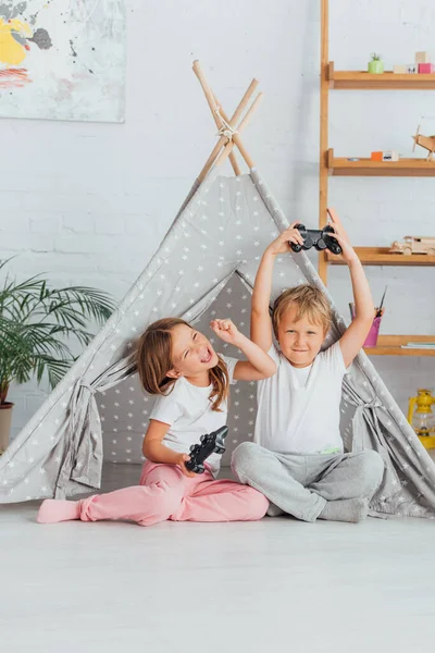 KYIV, UKRAINE - JULY 21, 2020: excited brother and sister in pajamas showing winner gesture while holding joysticks near kids wigwam — Stock Photo