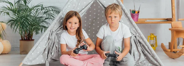 KYIV, UCRAINA - 21 LUGLIO 2020: immagine orizzontale di fratello e sorella in pigiama seduti sul pavimento vicino ai bambini wigwam e giocare ai videogiochi — Foto stock