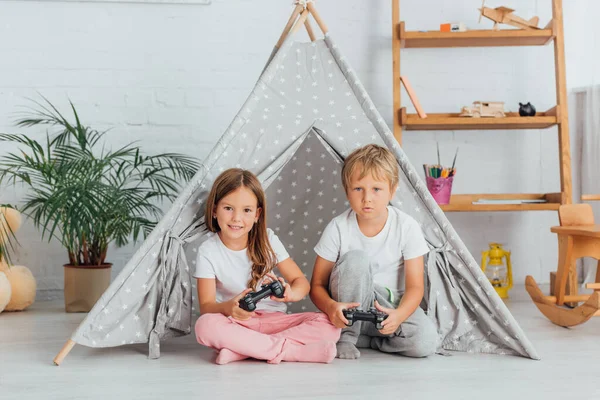 КИЕВ, Украина - 21 июля 2020 года: брат и сестра в пижаме сидят на полу рядом с игровой палаткой и играют в видеоигры — стоковое фото
