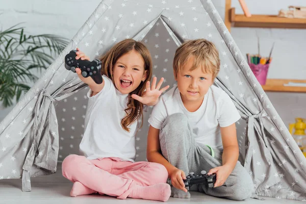 KYIV, UKRAINE - 21 juillet 2020 : fille excitée avec les mains tendues regardant la caméra tout en jouant au jeu vidéo avec frère près des enfants wigwam — Photo de stock