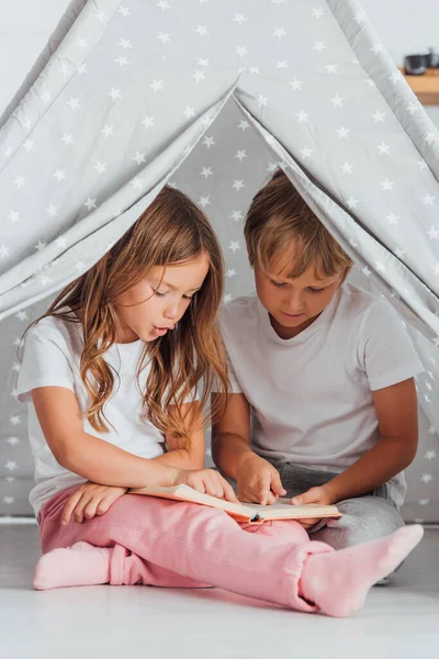 Hermana y hermano en pijama sentado en el suelo en tienda de juego y libro de lectura - foto de stock