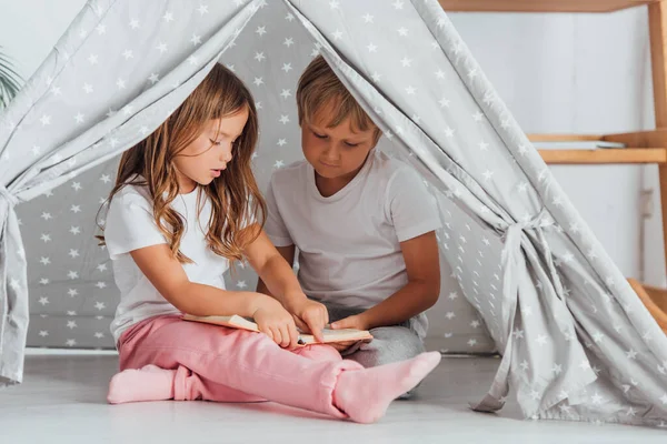 Fratello e sorella in pigiama seduti sul pavimento in bambini wigwam e libro di lettura — Foto stock