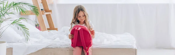 Imagen horizontal del niño en pijama sosteniendo la mochila escolar mientras está sentado en el dormitorio - foto de stock