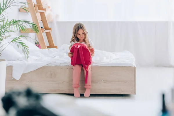 Selective focus of girl in pajamas holding school backpack while sitting on bed — Stock Photo