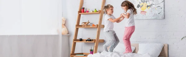 Imagen horizontal de hermano y hermana divirtiéndose tomados de la mano y saltando en la cama - foto de stock