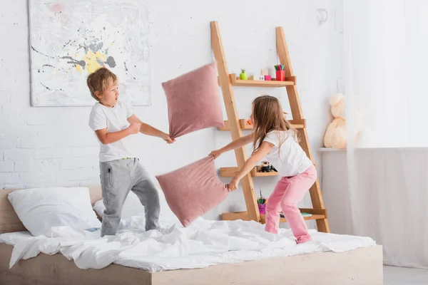 Сестра и брат в пижаме дерутся с подушками, веселясь в спальне — стоковое фото