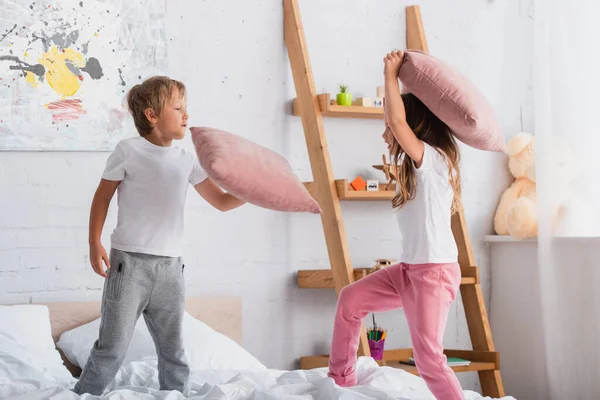 Sister and brother in pajamas having fun while fighting with pillows in bedroom — Stock Photo