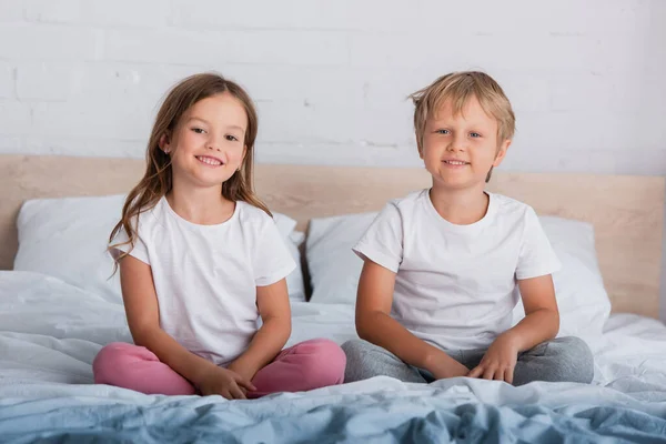 Brother and sister in pajamas looking at camera while sitting with crossed legs on bed — Stock Photo