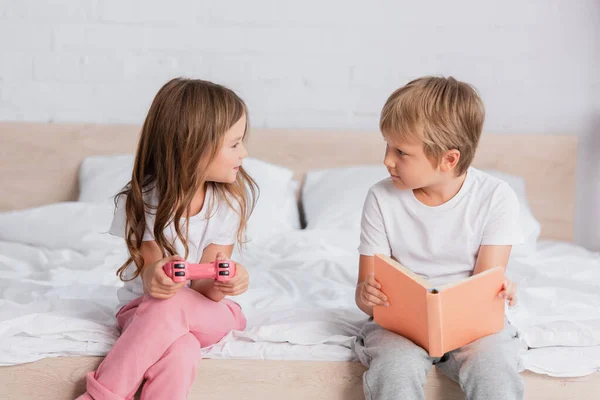 KYIV, UKRAINE - JULY 21, 2020: girl with joystick and boy with book looking at each other while sitting on bed — Stock Photo