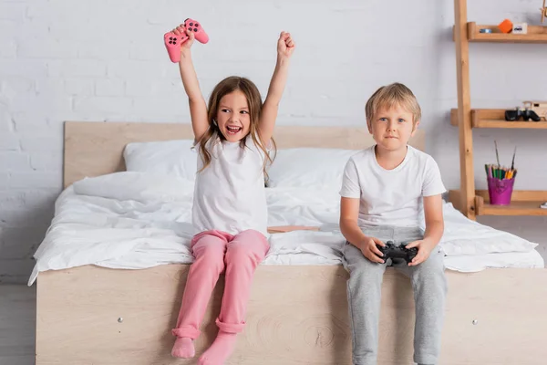 KYIV, UKRAINE - JULY 21, 2020: excited girl showing winner gesture near brother holding joystick in bedroom — Stock Photo