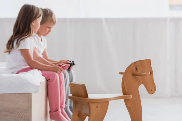 KYIV, UCRANIA - 21 de julio de 2020: enfoque selectivo del hermano y la hermana en pijamas jugando videojuegos cerca del caballo mecedora - foto de stock