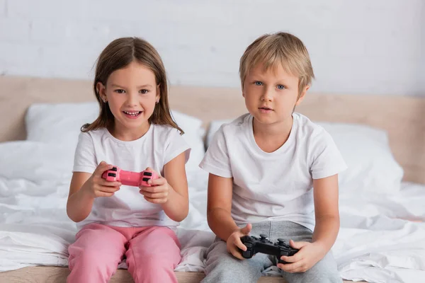 KYIV, UCRANIA - 21 de julio de 2020: chica emocionada y hermano concentrado en pijamas jugando videojuegos en el dormitorio - foto de stock
