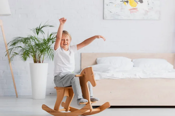 Excited kid riding rocking horse with raised hands near potted plant and bed — Stock Photo
