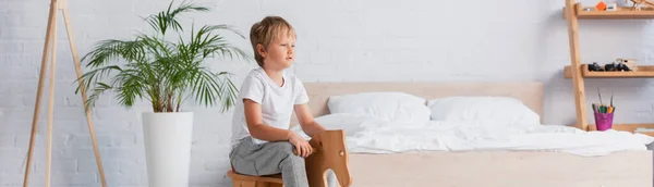 Horizontal image of boy in pajamas riding rocking horse near potted plant and bed — Stock Photo