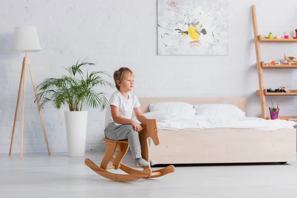 Boy in pajamas riding rocking horse near potted plant and bed in spacious bedroom — Stock Photo