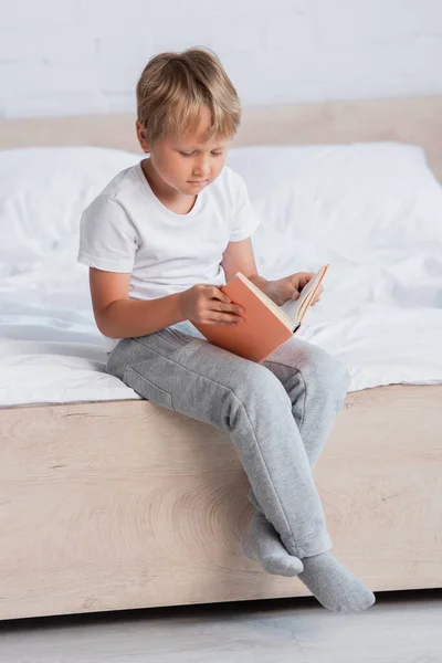 Libro de lectura infantil concentrado mientras está sentado en la cama en pijama - foto de stock