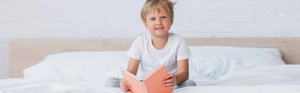Concepto horizontal de niño en camiseta blanca mirando a la cámara mientras está sentado en la cama con libro - foto de stock