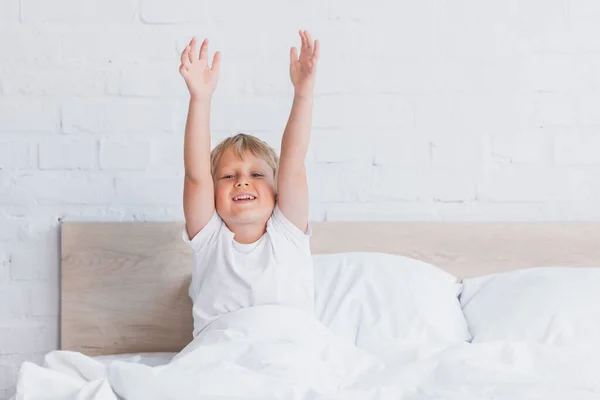 Пробуджений хлопчик у білій футболці дивиться на камеру, розтягуючись у ліжку — стокове фото