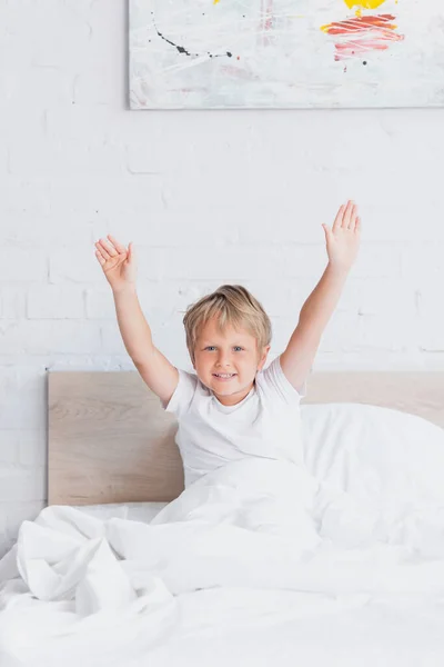 Garçon réveillé en t-shirt blanc étirant tout en étant assis dans le lit — Photo de stock